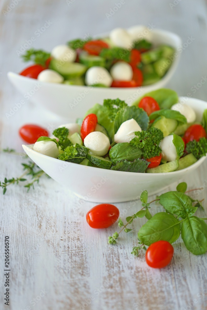Vegetable summer salad from tomatoes, cucumbers, mozzarella, parsley, basil in white cups on a white shabby wooden background.Vegetarian menu.	