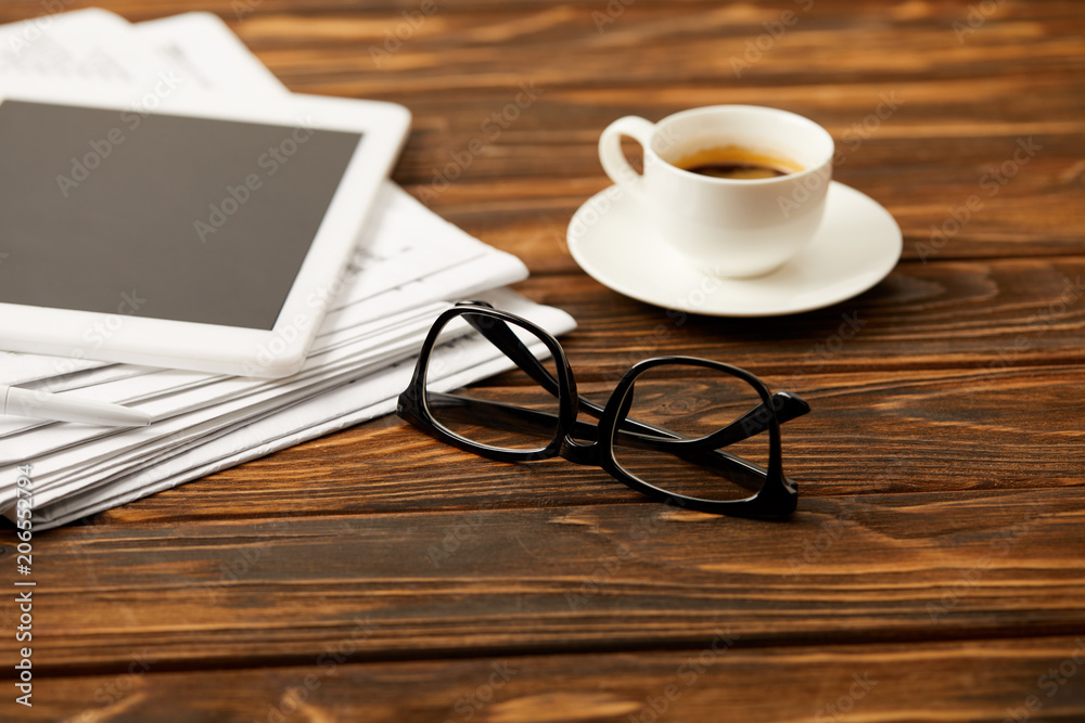 cup of coffee eyewear, digital tablet and pile of newspapers on wooden background