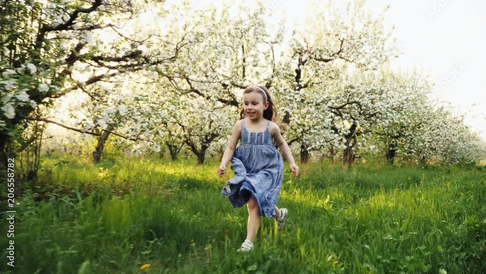 Slow motion little brunette girl with blue dress running in exterior garden green grass and plants Slow motion little brunette girl with blue dress running in exterior garden green grass and plants