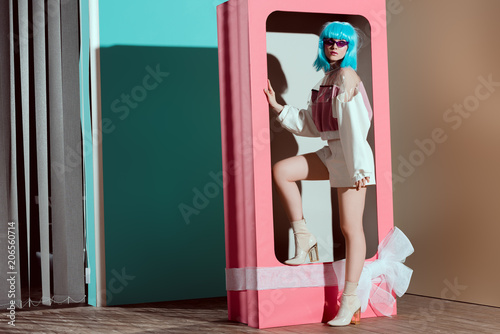 fashionable young woman in blue wig posing in decorative pink box with bow