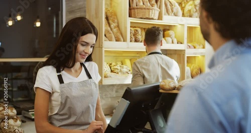 Young beautiful woman standing at the counter in the bakery shop and selling bread to the man, her male co-worker doing something behind her. Indoors photo