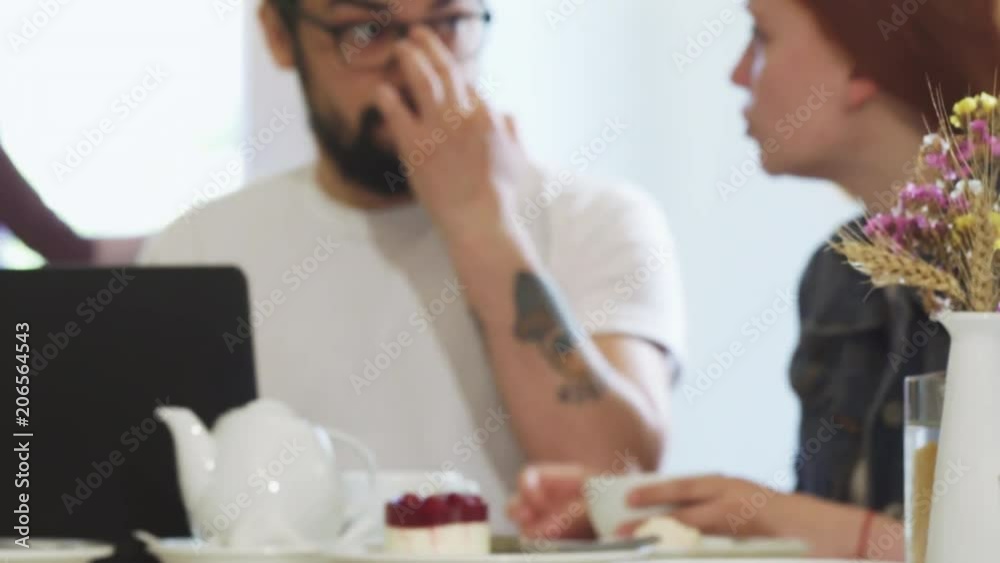 Sliding shot of a young couple having breakfast together at the coffee shop. Handsome bearded man using laptop and talking to his beautiful girlfriend. Communication, dating.