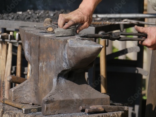 steel anvil, on top of which lies a hot workpiece which is held by a pair of pliers. The blacksmith holds the pliers with one hand, with the other it processes the workpiece, further hammers visible. 