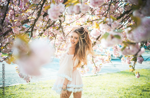 Girl in short pink dress enjoying sunny day in botanical garden. Female playing with long gorgeous blond hair, beauty and hair care products. Sexy woman posing under blooming cherry blossom tree