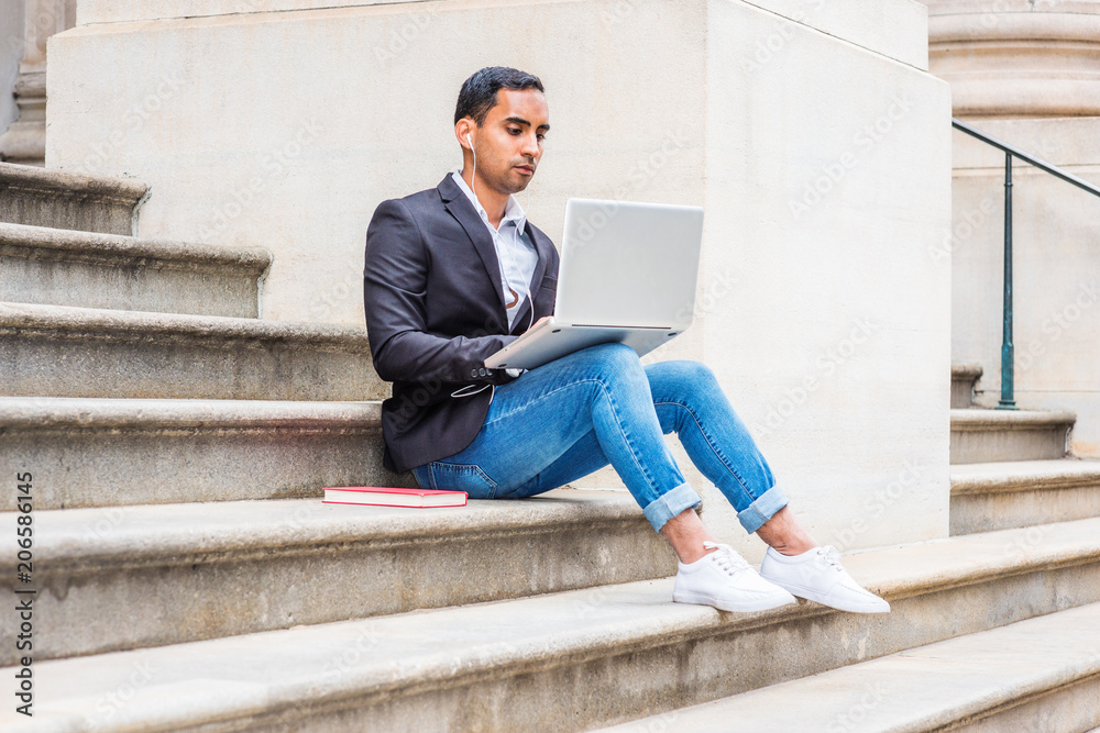 Young Hispanic American college student studying in New York, wearing black blazer, blue jeans, sneakers, earphone, sitting on stairs of office building, listening music, working on laptop computer..