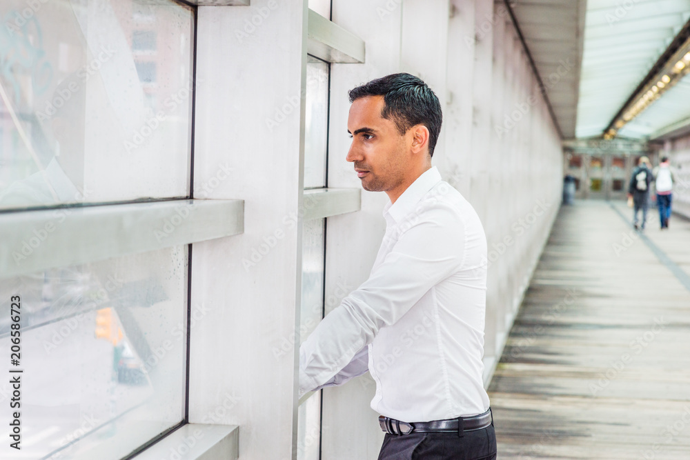 Young Handsome Hispanic American Businessman working in New York, wearing white shirt, black pants, standing on walkway by glass wall inside office building, looking outside, thinking, lost in thought