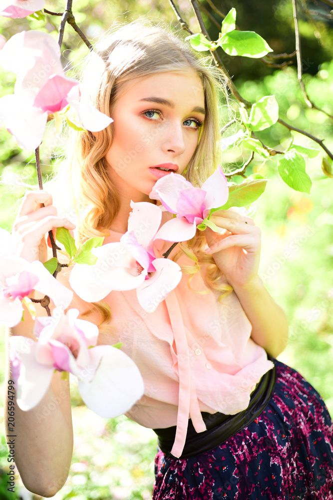Fototapeta premium Charming blond girl enjoying scent of flowering trees. Pretty lady relaxing and breathing fresh aroma of blooming magnolia. Attractive curly-haired young woman walking through the spring garden