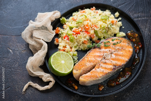 Grilled salmon fillet and cuscus with vegetables on a metal tray, studio shot on a brown stone background