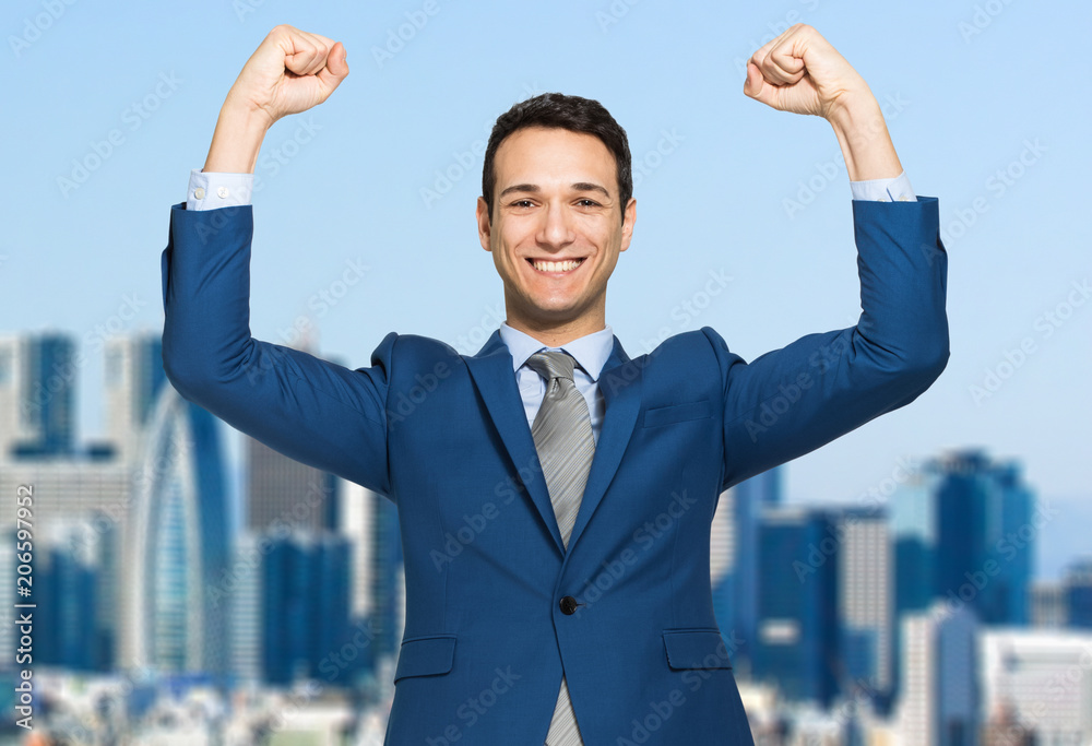 Businessman smiling and raising his fist in the air, with office building in background - business success, achievement, and win concepts Businessman smiling and raising his fist in the air, with office building in background - business success, achievement, and win concepts
