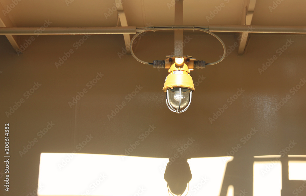 Yellow Lamp on Ship Deck Surrounded by Metal Rustic Frame Fixed to a Ceiling. Bulkhead Light on Navy Ship against Vintage Painted Wall Background. Industrial Light Bulb Lamp Detail Close Up View.