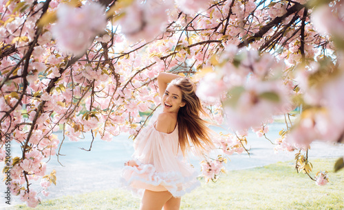 Woman with gorgeous smile having fun in blooming garden. Female with slim sexy body on natural background, wellness and fitness concept. Sensual blond girl in short chiffon dress walking in park
