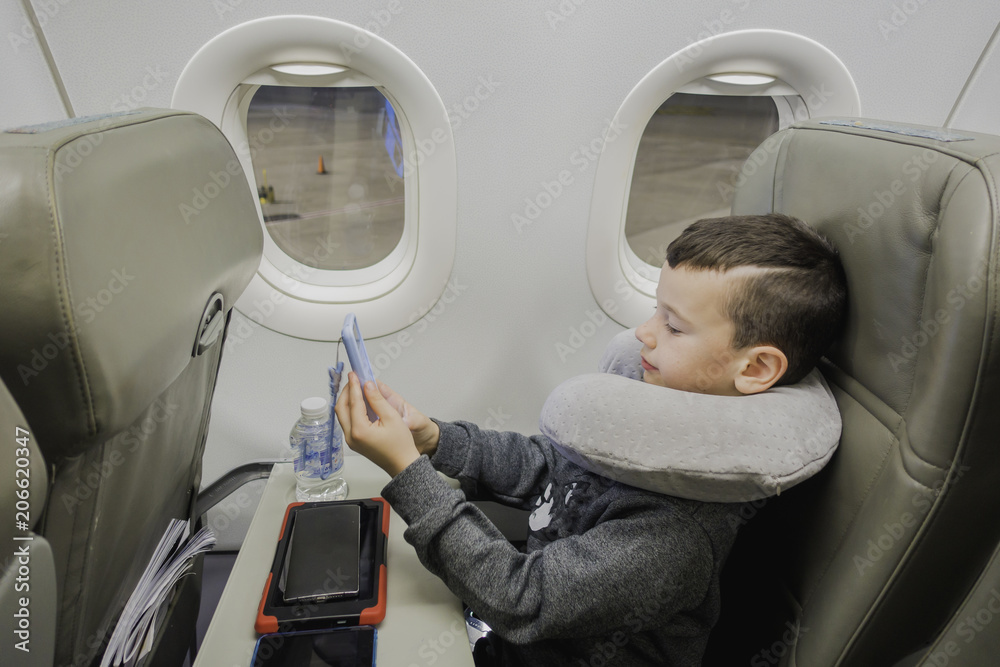 A boy is sitting in a plane near the porthole With travel pillow ,playing in a gadget and waiting for take-off. A boy is sitting in a plane near the porthole With travel pillow ,playing in a gadget and waiting for take-off.