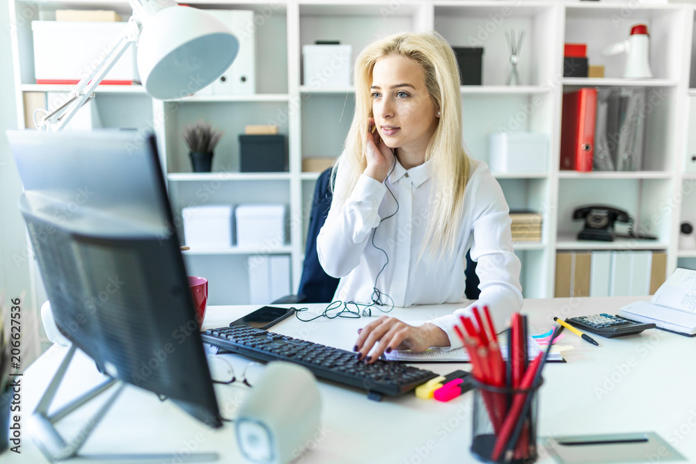 A young girl in the office sits at a table talking on the phone through a headset and working at the computer. A young girl in the office sits at a table talking on the phone through a headset and working at the computer.