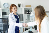 A beautiful young girl in a white coat is standing near the desk in the office and shaking hands with the patient. The patient thanks the doctor.