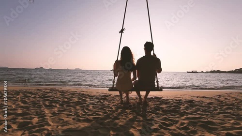 romantic couple in love sitting together on rope swing at sunset beach, silhouettes of young man and woman on holidays or honeymoon