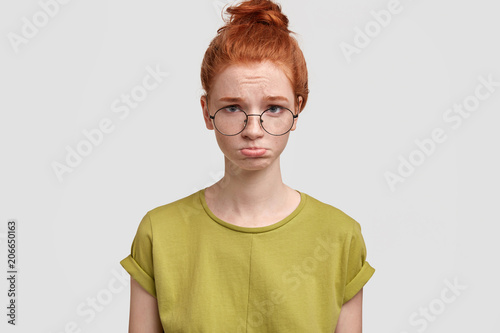 Portrait of discontent female frowns face in discontent, looks desperately at camera, going to cry, has freckled skin, poses against white studio wall. Redhead woman cries and being offended