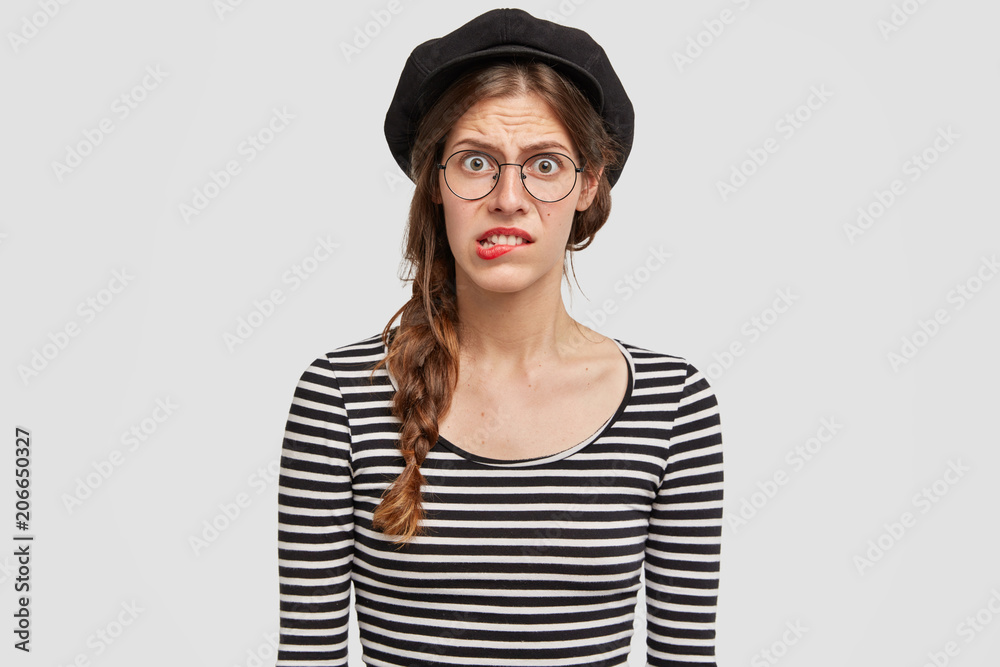 Puzzled nervous French woman bites lips nervously, wears beret and striped jacket, feels frustration, poses against white background. Parisian female has some troubles, sees no way out, worries Puzzled nervous French woman bites lips nervously, wears beret and striped jacket, feels frustration, poses against white background. Parisian female has some troubles, sees no way out, worries