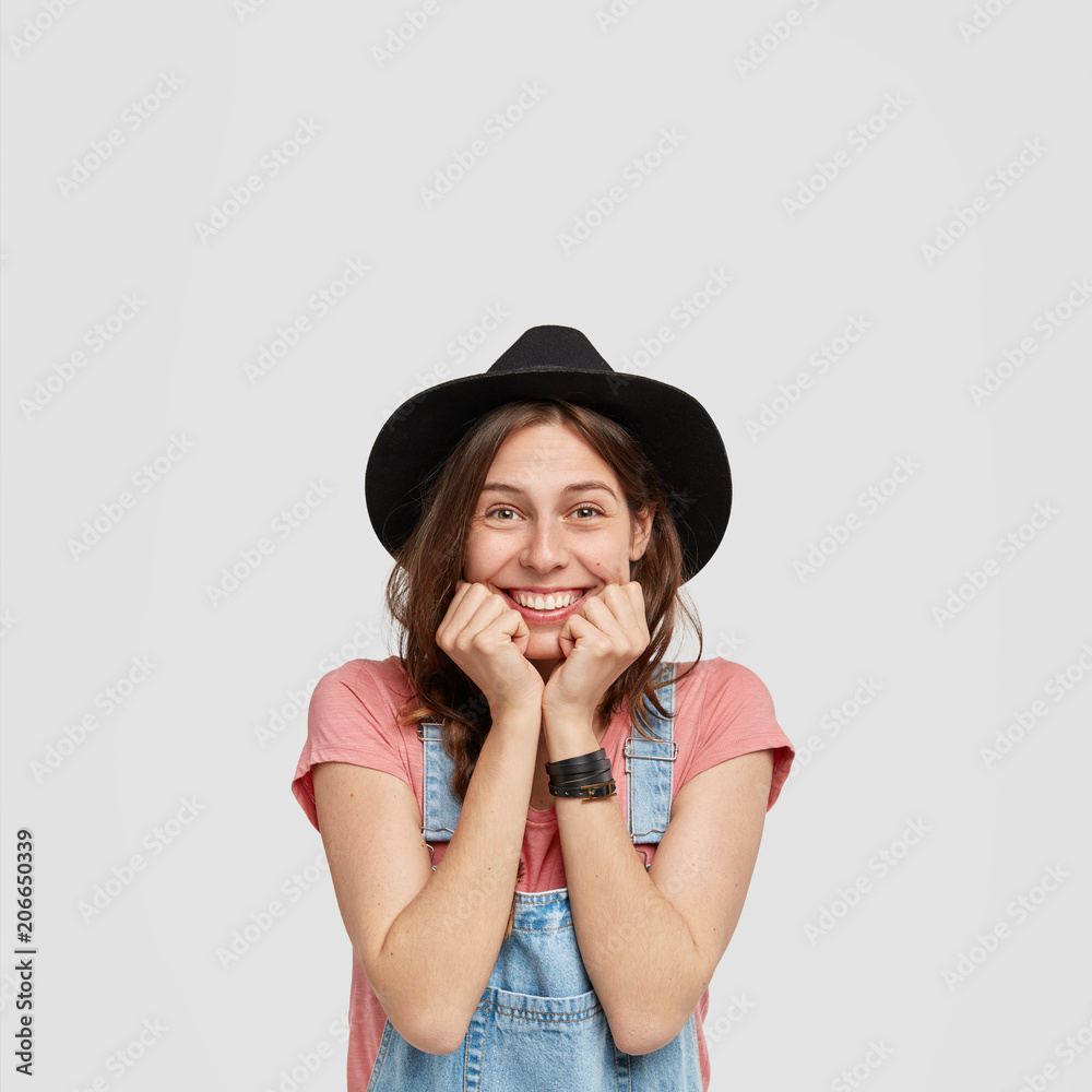 Vertical shot of happy young female engaged in agriculture, keeps hands under chin, hears good comments about her garden, has positive expression, wears casual clothes, isolated over studio wall