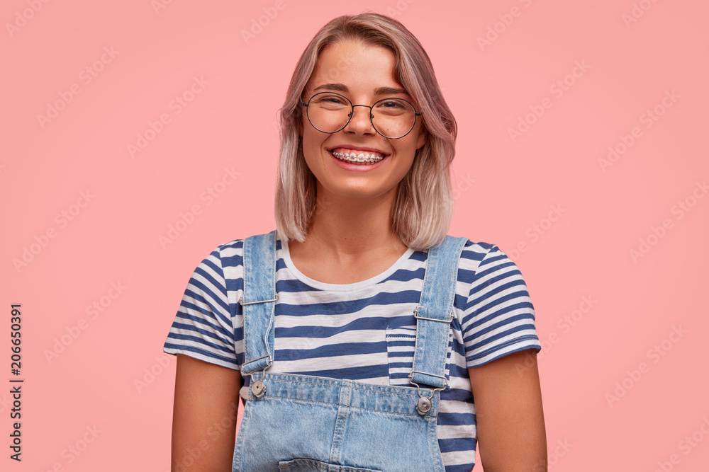 Obraz premium Glad smiling pretty female freelancer, has broad smile, demonstrates teeth with braces, wears casual t shirt with overalls, rejoices recieving reward, stands against pink background. Emotions concept