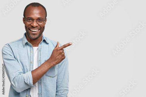Happy delighted male manager involved in advertising company`s product, indicates with fore finger at blank copy space, wears denim shirt, has positive smile. Advertisement and ethnicity concept