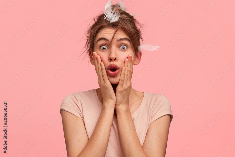 Omg, I overslept! Stunned beautiful young female keeps hand on cheeks and looks with shocked expressin, wakes up late in morning, has feathers on head, wears casual outfit. Awakening concept Omg, I overslept! Stunned beautiful young female keeps hand on cheeks and looks with shocked expressin, wakes up late in morning, has feathers on head, wears casual outfit. Awakening concept