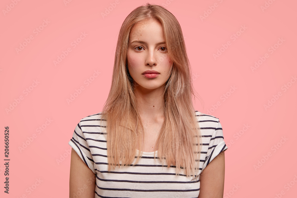 Studio shot of serious young European female with light hair, dressed in striped t shirt, thinks about future plans, stands against pink background. Beautiful woman stands indoor. Beauty concept Studio shot of serious young European female with light hair, dressed in striped t shirt, thinks about future plans, stands against pink background. Beautiful woman stands indoor. Beauty concept