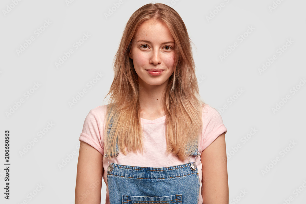 Attractive teenager with confident look, has long straight hair, dressed casually, smiles gently, looks directly at camera, reads necessary information in front, stands against white concrete wall