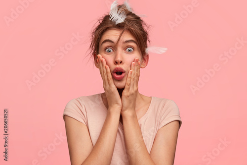 Omg, I overslept! Stunned beautiful young female keeps hand on cheeks and looks with shocked expressin, wakes up late in morning, has feathers on head, wears casual outfit. Awakening concept photo
