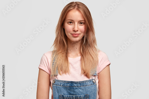 Attractive teenager with confident look, has long straight hair, dressed casually, smiles gently, looks directly at camera, reads necessary information in front, stands against white concrete wall © wayhome.studio 