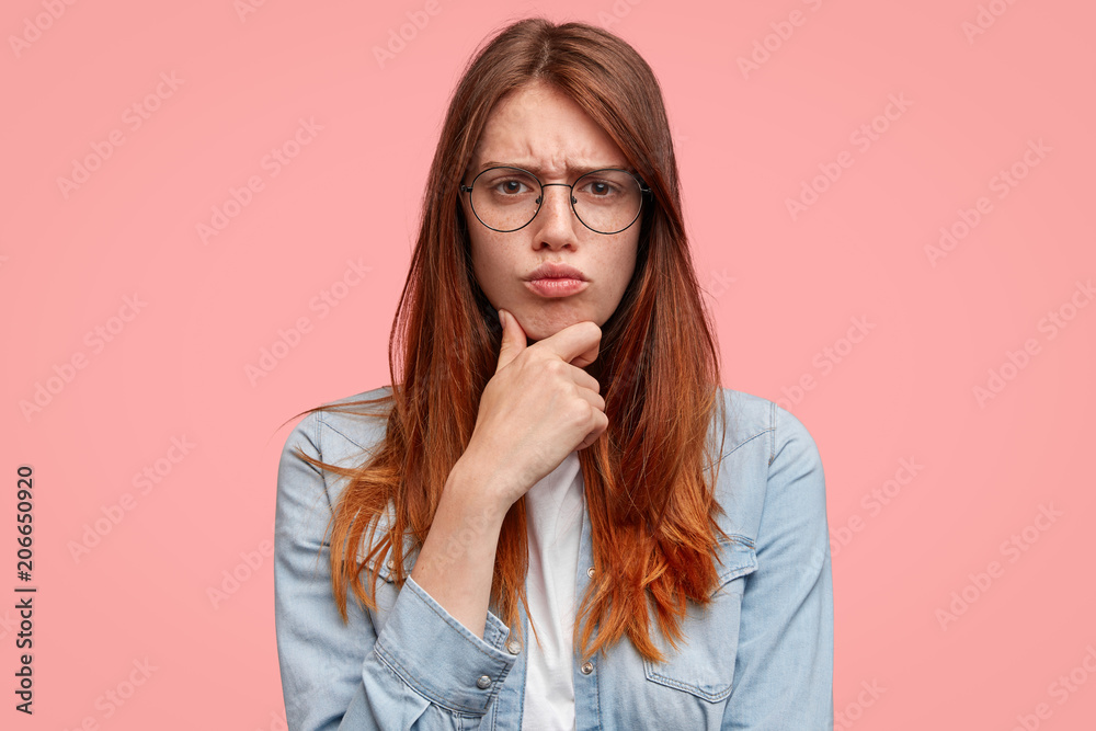 I tell definitely no. Gloomy and discontent female with freckled face, holds chin and frowns face, being in bad mood, makes displeased grimace, wears denim jacket, isolated on pink background I tell definitely no. Gloomy and discontent female with freckled face, holds chin and frowns face, being in bad mood, makes displeased grimace, wears denim jacket, isolated on pink background