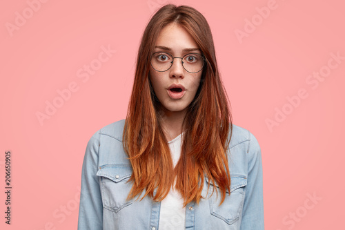 Amazed attractive freckled female with wide opeed mouth, has straight hair and freckled skin, wears round spectacles and fashionable denim jacket, being afraid of something. Stunned student at college