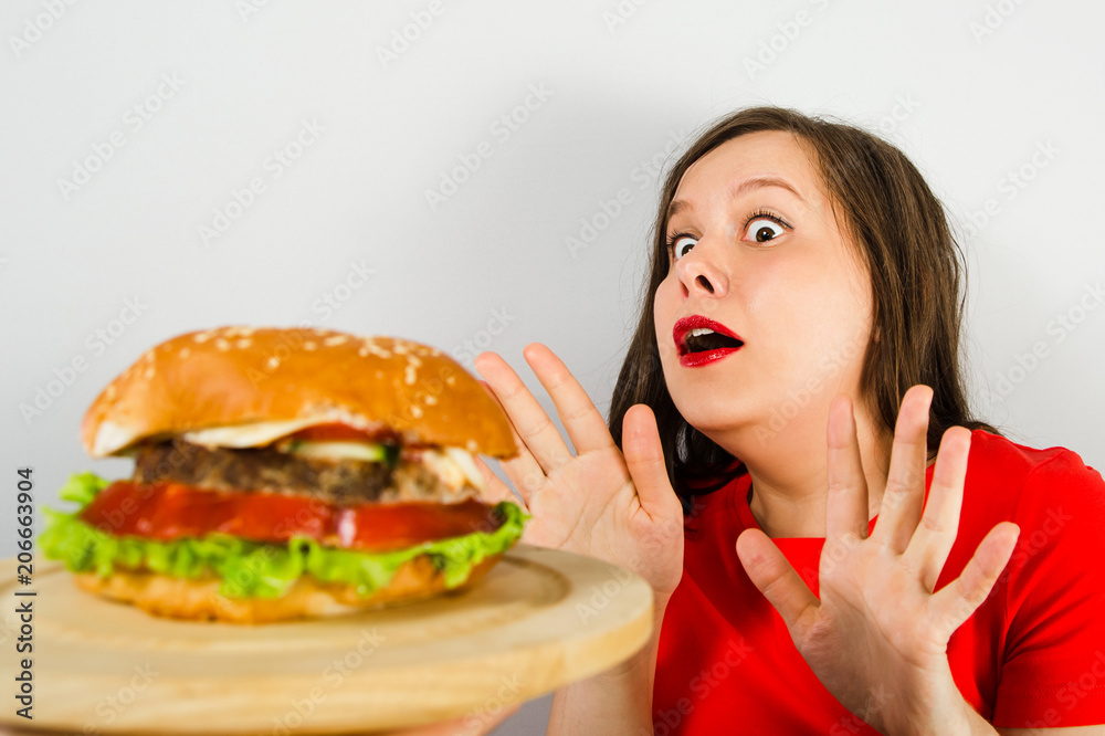 Young fat woman stretched out her hands for fear of a hamburger on gray background. Young fat woman stretched out her hands for fear of a hamburger on gray background.