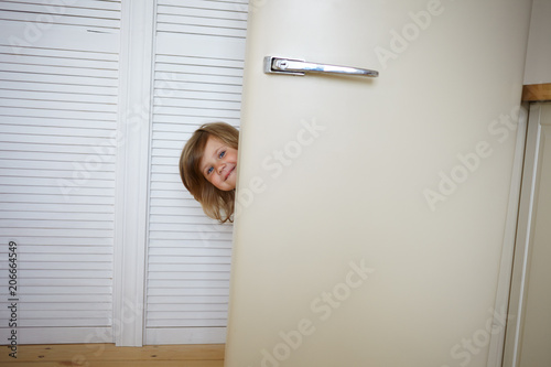 Indoor shot of funny adorable European little 5 year old girl playing hide and seek in modern kitchen interior, hiding herself behind fridge door. Childhood, fun, joy, games and happiness concept