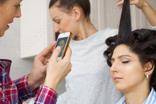 Salon master taking photo on smartphone of beauty woman while putting make-up on and making hairdro. photo