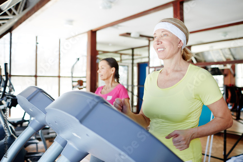 Happy aged woman running on treadmill in sports center with her friend on background