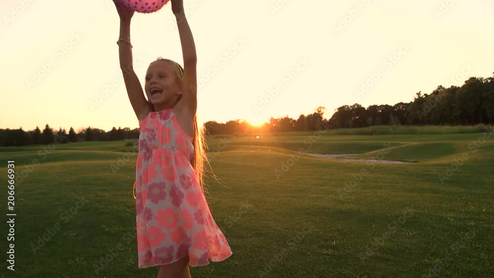 Cute girl caught the ball outdoors. Little girl in pink dress having fun with ball on green lawn at sunset, slow motion. Beauty of nature and childhood.