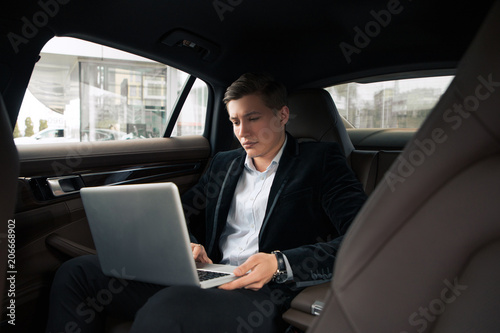 It is time to work! Handsome good-looking young busy man seriously doing his work on the laptop on the rear seat in the car.
