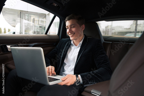Work with pleasure! Handsome smiling young man doing the business on the back seat of his car, while using the wireless computer. © stacestock