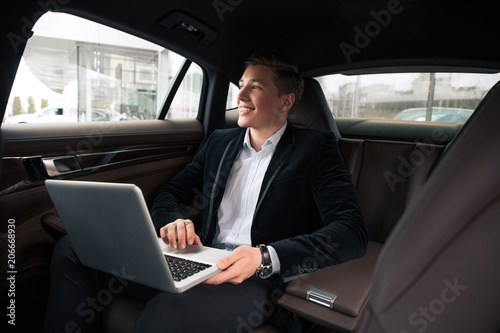 Cheerful work! Smart clever young smiling newcomer businessman using internet, while working with laptop in the automobile. © stacestock