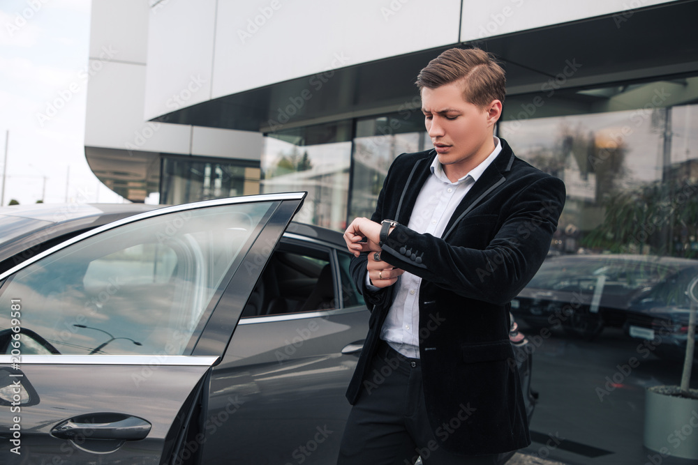 Do not hurry up! Attractive intelligent young businessman checking the time, while standing and waiting for someone near the automobile. Do not hurry up! Attractive intelligent young businessman checking the time, while standing and waiting for someone near the automobile.