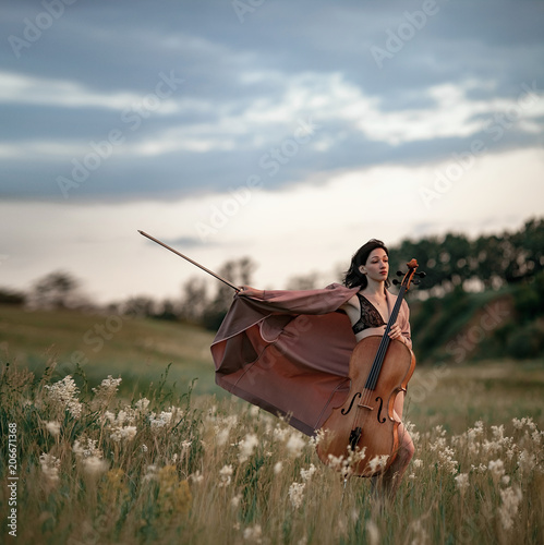 Woman cellist in unbuttoned cloak prepares to play at flowering meadow against backdrop of picturesque landscape. photo