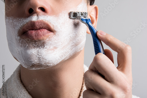 No stubble. Close up half of face with foam of young man who is standing and shaving his cheeks. He is holding blue razor and carrying about his appearance. Male beauty concept