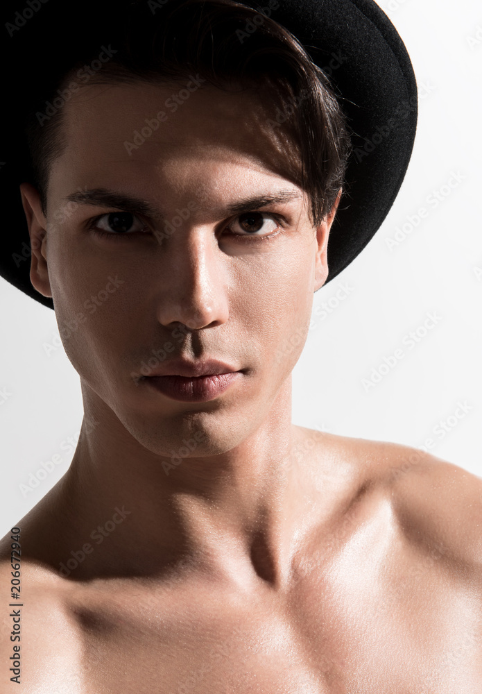Portrait of naked young male with clean and healthy skin in stylish hat is standing against light background and looking at camera decisively. Masculine style and beauty concept