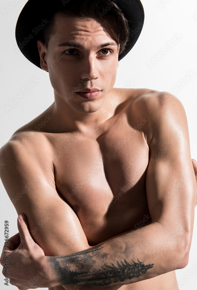 Portrait of topless cute guy in stylish hat is standing against light wall. He looking forward dreamily with tattooed arms crossed on chest. Masculine beauty and style concept Portrait of topless cute guy in stylish hat is standing against light wall. He looking forward dreamily with tattooed arms crossed on chest. Masculine beauty and style concept