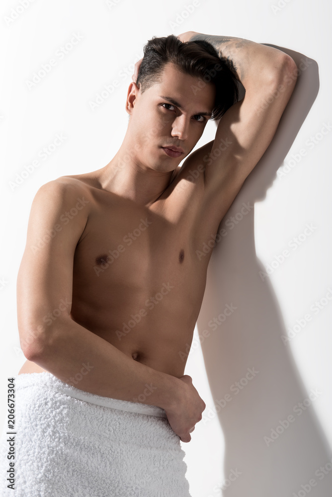 Fototapeta premium Hopeless. Portrait of shirtless youthful man with trained gorgeous body is keeping head by hand while leaning on white wall. He is standing with towel on his hips and looking at camera thoughtfully