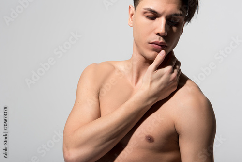 Sensitive appearance. Calm young attractive naked man is standing against light background and touching his neck. He is looking down dreamily. Copy space in the left side. Soft skin concept