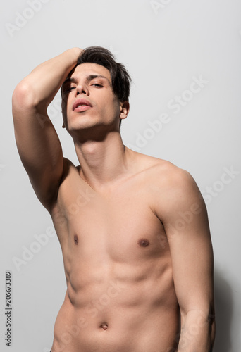 Perfect male body. Low angle portrait of shirtless cute guy is standing against light wall and looking at camera thoughtfully. He is posing with raised hand. Masculine beauty concept