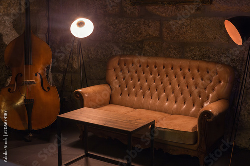 Modern jazz bar interior design, leather sofa, cello music instrument, lamps. Stone wall on background, brutal atmosphere
