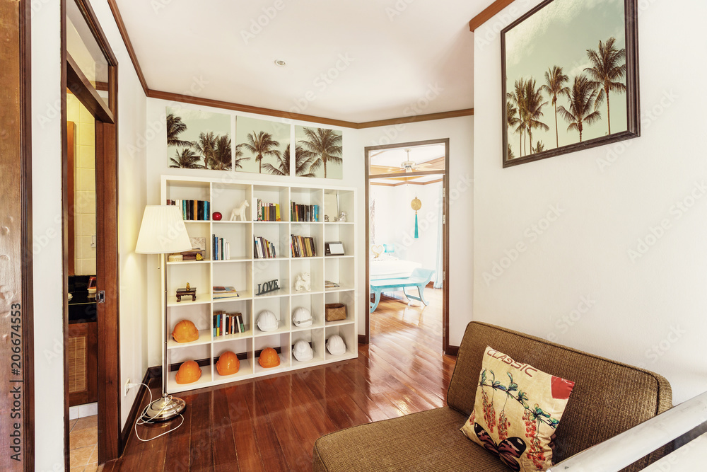 Fototapeta premium Small living room interior. Shelfs with books and construction helmets like decor elements, tropical palm trees in pictures on the wall, open doors to bedrooms
