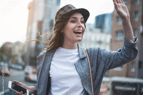 Attractive smiling lady is raising arm to say hello. She is wearing trendy hat and holding smartphone in the street